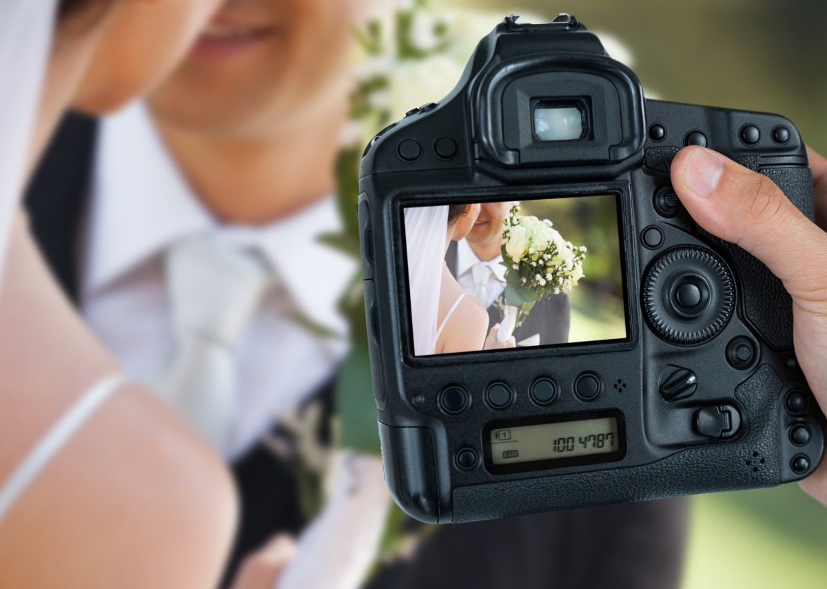 Someone holds a camera in front of a bride and groom posing. The bride's bouquet is visible through the camera lens.