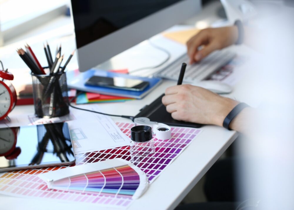 A person using a stylus beside printed color swatches, ink jars, pencils, a tablet, and a fan deck on a table.