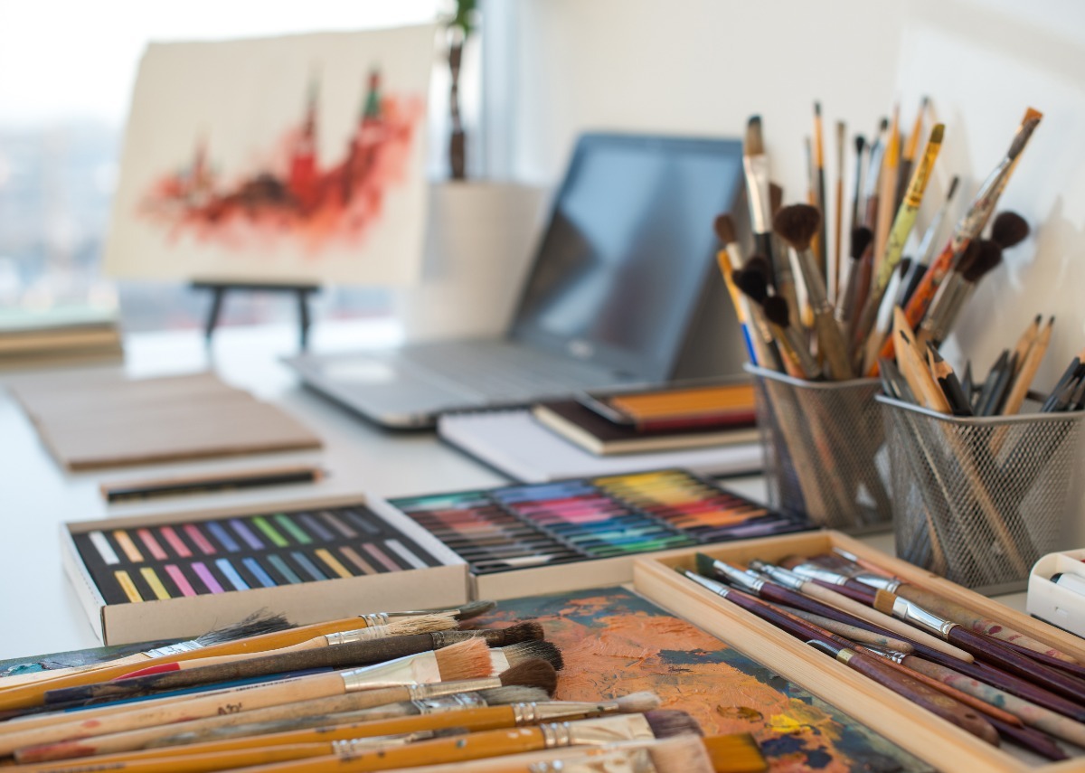 A desktop with cups of brushes and pencils, pastel trays, a laptop, and an abstract canvas near a window.